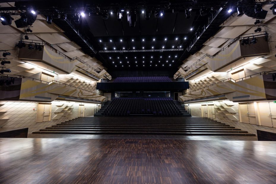 Das Bild zeigt eine moderne Theater- oder Veranstaltungsstätte in einem weitläufigen Innenraum. Der Blick ist vom Bühnenbereich aus auf die Zuschauerränge gerichtet. Die Sitzreihen sind in einer steilen Anordnung angeordnet, was eine gute Sicht auf die Bühne ermöglicht. Das Design ist minimalistisch und elegant, mit hellen Wänden und einem dunklen Parkettboden. Über der Bühne hängen zahlreiche Scheinwerfer und Lichtquellen, die eine intensive Beleuchtung ermöglichen. Die gesamte Atmosphäre wirkt einladend und funktional, ideal für verschiedene kulturelle Veranstaltungen oder Aufführungen.