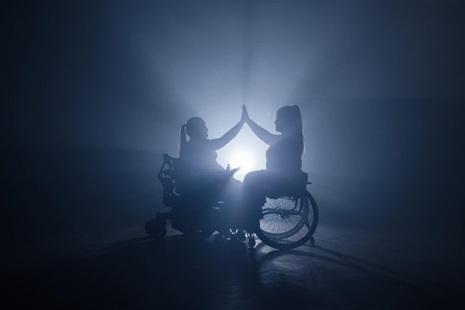 Das Bild zeigt zwei Frauen in Rollstühlen, die sich gegenüberstehen und mit ihren Handflächen in einer symbolischen Geste der Verbundenheit oder des Einvernehmens Hände schütteln. Es handelt sich um Silhouetten, die vor einem hellen, diffusen Licht stehen, das eine dramatische Atmosphäre schafft. Diese Darstellung vermittelt ein Gefühl von Gemeinschaft und Solidarität, während sie gleichzeitig die Stärke und den Mut der dargestellten Personen betont. Der dunkle Hintergrund hebt die beiden Frauen hervor und lenkt die Aufmerksamkeit auf ihren Akt der Interaktion.