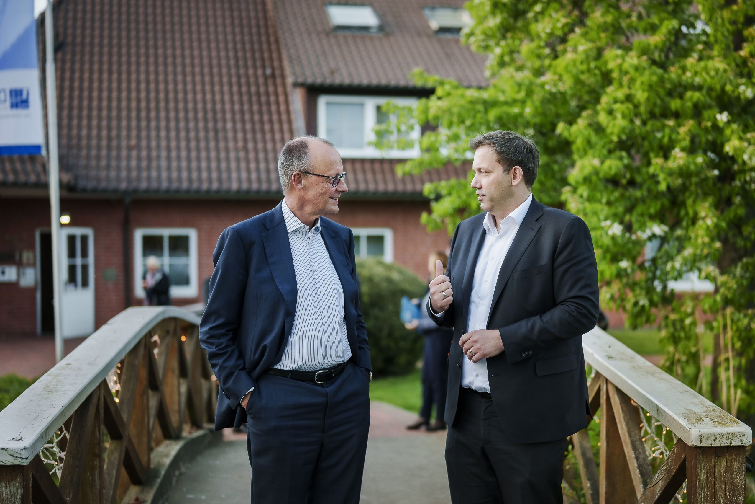 Zwei Männer im Anzug stehen auf einer kleinen Holzbrücke und unterhalten sich. Im Hintergrund befinden sich ein Gebäude und ein grüner Garten. Das Bild vermittelt einen politischen Austausch in ruhiger Atmosphäre.